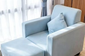 Ein hellblauer Sessel mit passendem Hocker in einem Raum mit Holzboden und einem Fenster mit durchsichtigen Vorhängen.