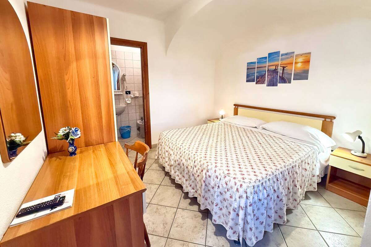 Un dormitorio con un escritorio de madera, una cama con colcha estampada floral, una mesita de noche con lámpara y una pared decorada con tres cuadros de paisajes al atardecer.