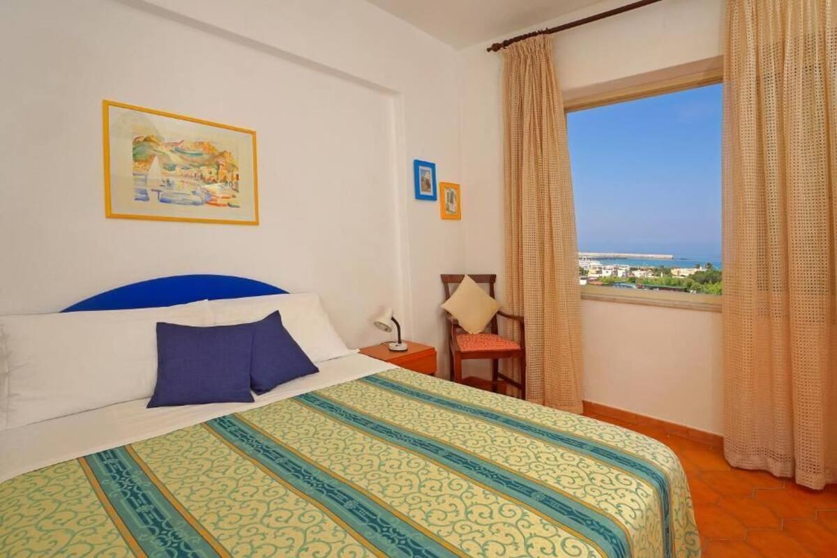 Una habitación de hotel con cama, una silla, una mesita de noche, una ventana con cortinas y un cuadro en la pared.