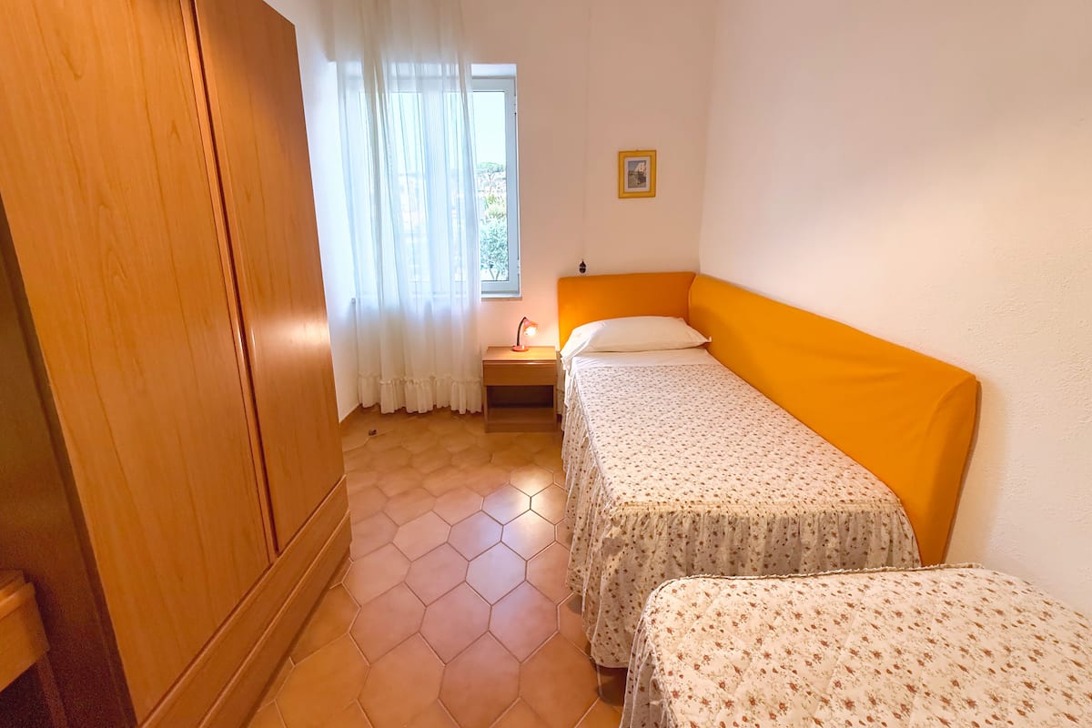 Una habitación con dos camas individuales, un armario de madera, un ventanal con cortinas translúcidas y una mesita de noche con lámpara.