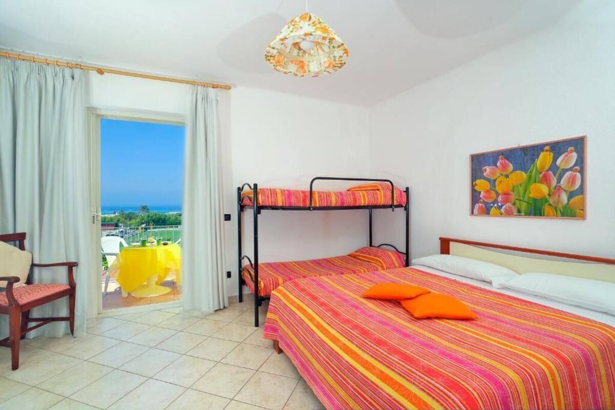 Habitación de hotel con una cama nido, una cama individual, un cuadro de tulipanes, un balcón con mesa amarilla y vista al mar.