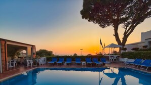 Una piscina con sillas reclinables azules y vistas al atardecer.