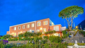 Un hotel de varios pisos con una fachada cálida, un árbol grande al frente y un muro de piedra.
