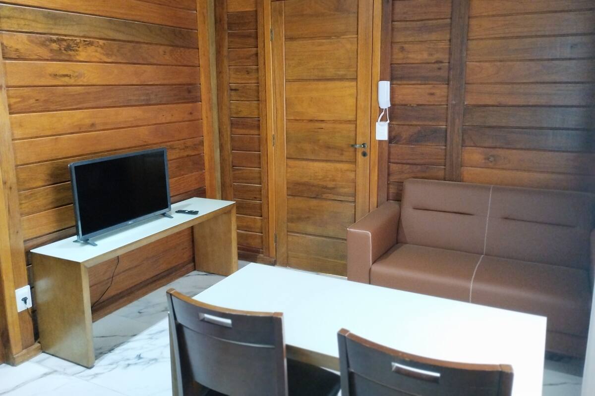 Une pièce aux murs lambrissés, avec un bureau blanc, un téléviseur, un canapé marron et deux fauteuils.