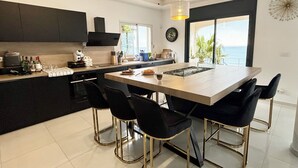 A modern kitchen with a central island, black bar stools, and a large window offering a view of the ocean. - Maison Paradisiaque A Rosas A Proximite DES Plages (CANYELLES ALMADRABA (AJUNTAMENT ROSES))