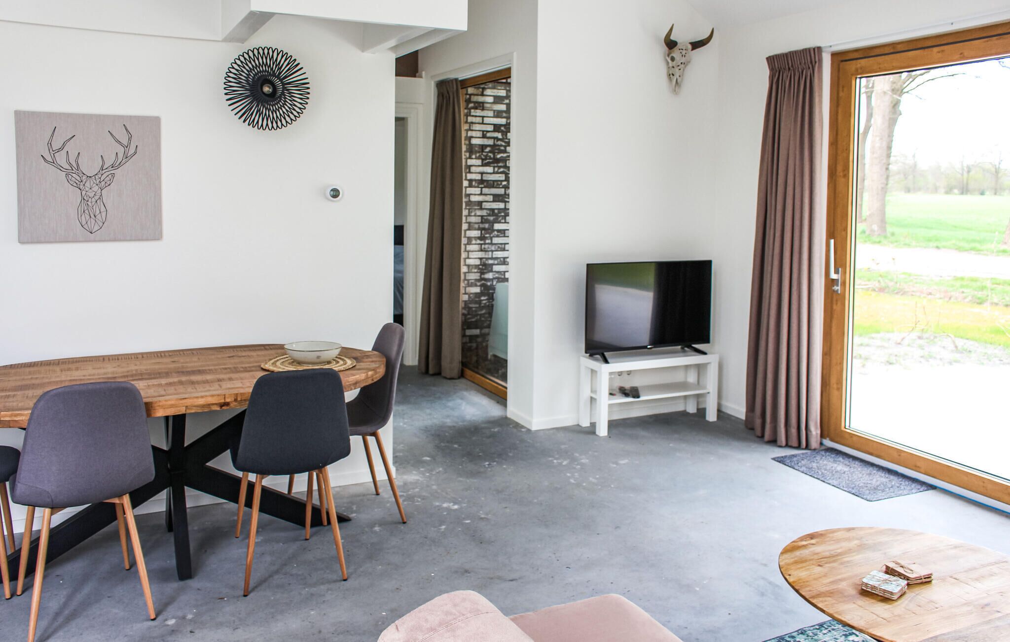 A modern living room with a wooden dining table, two grey chairs, a TV on a white stand, and a stone wall.