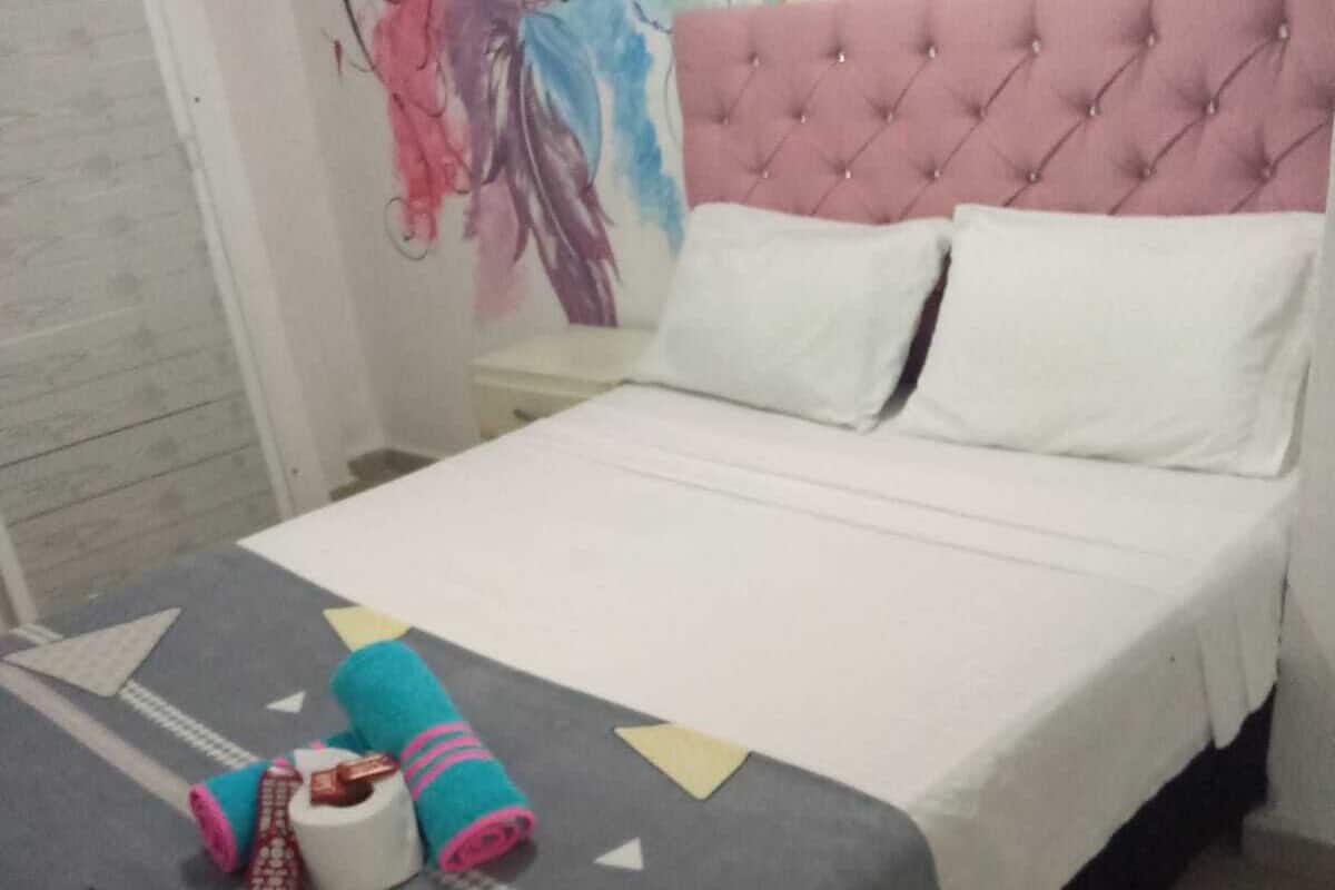 Una cama impecable con un cabecero rosa con capitoné, almohadas blancas y una colcha gris con patrones geométricos y la palabra 'love'.