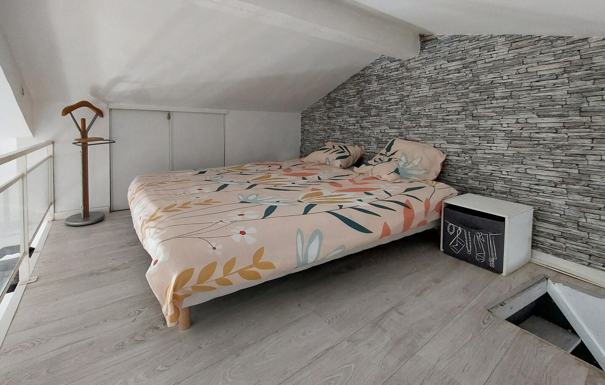 A bedroom with a bed, a nightstand, a wooden floor, a stone wall, and a window.