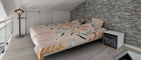 A bedroom with a bed, a nightstand, a wooden floor, a stone wall, and a window.