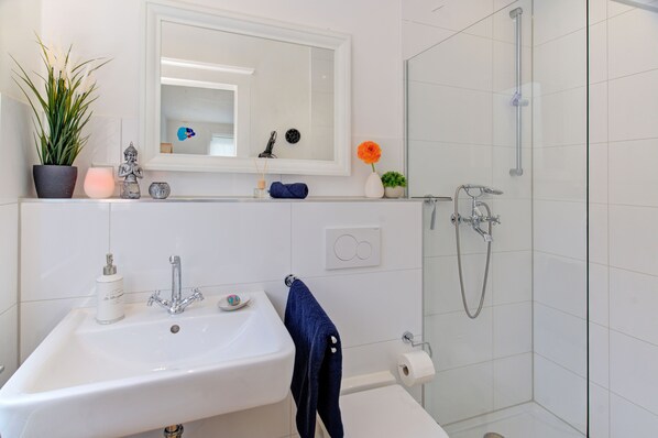 A bathroom with a white sink, mirror, and shower. There is a potted plant, a candle, and a small statue on the counter.