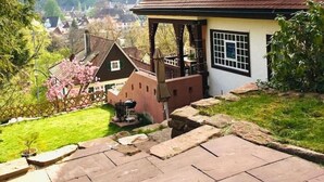Una casa a due piani con tetto spiovente, un portico e vista su un villaggio sullo sfondo.