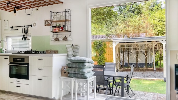 Cocina moderna con armarios blancos, horno empotrado y comedor con mesa y sillas. Se ve un jardín y árboles por la ventana.