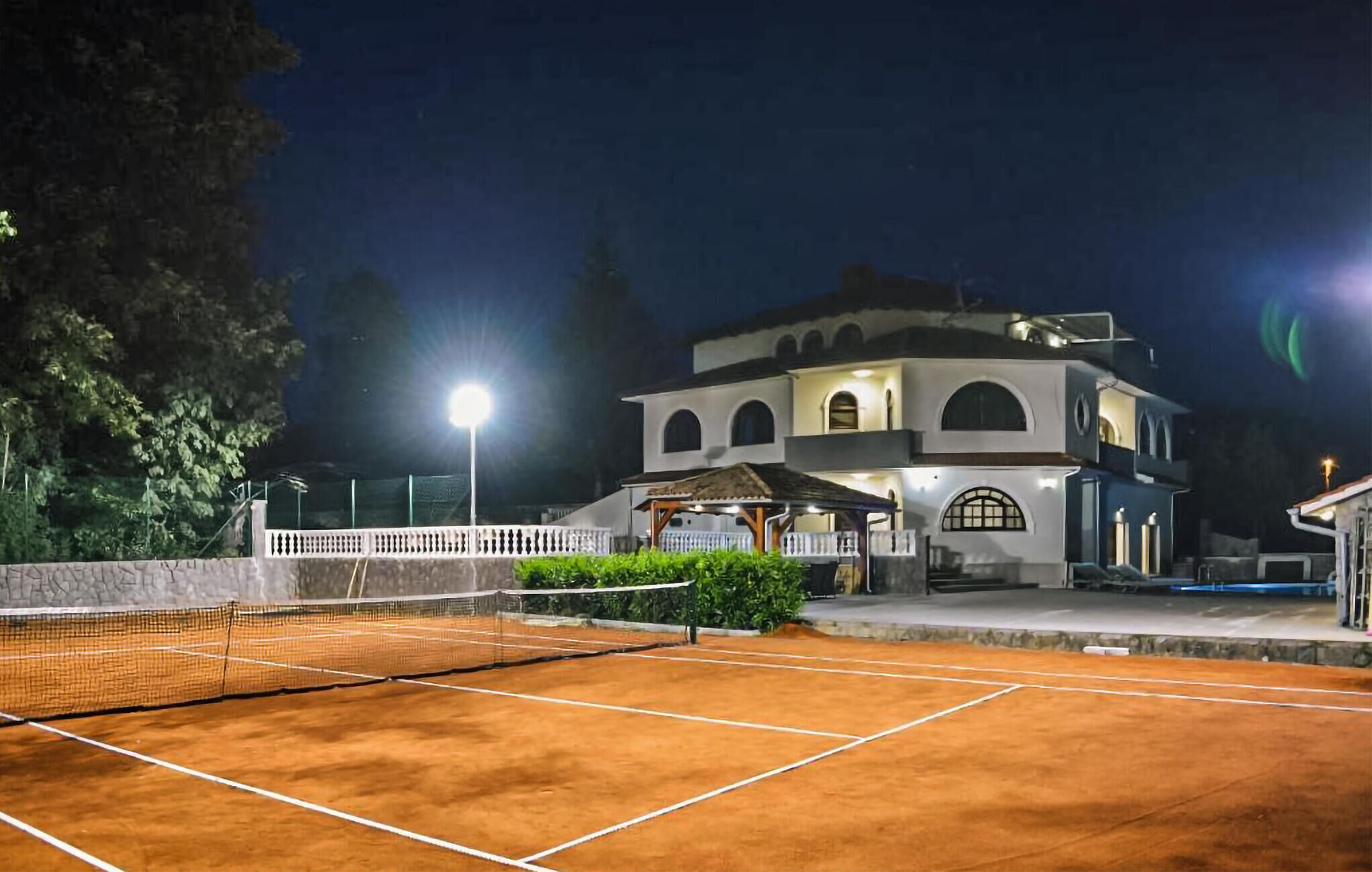 A tennis court with a net, surrounded by a white fence and a building with multiple arches and a covered seating area.