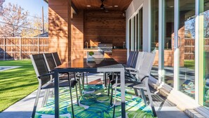 Eine Terrasse im Freien mit Holzboden, einer Glastür, die zu einer Küche führt, und einem Esstisch mit Stühlen.