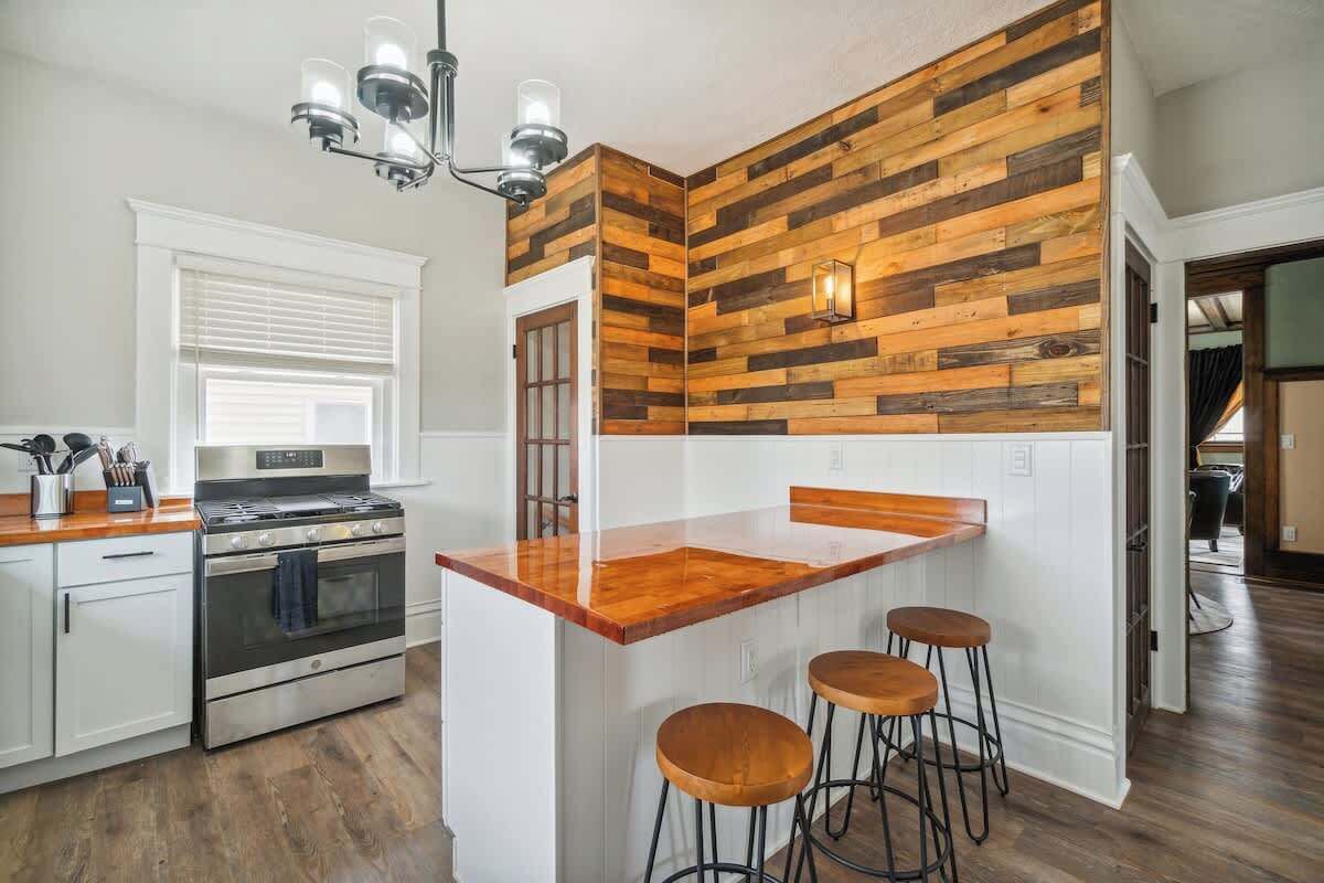 A kitchen with a wooden accent wall, a stainless steel oven, a wooden countertop, and bar stools.