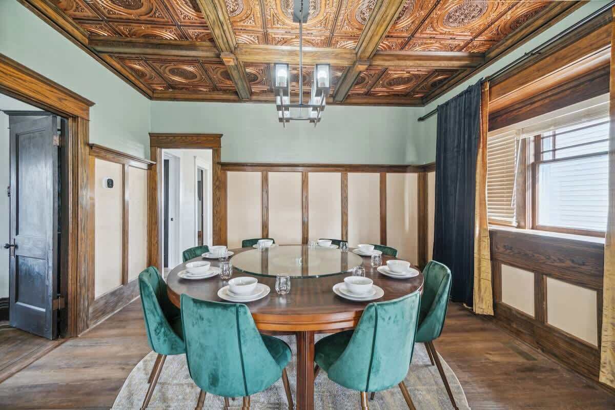A dining room with a round table, green upholstered chairs, and a wooden ceiling with intricate designs.