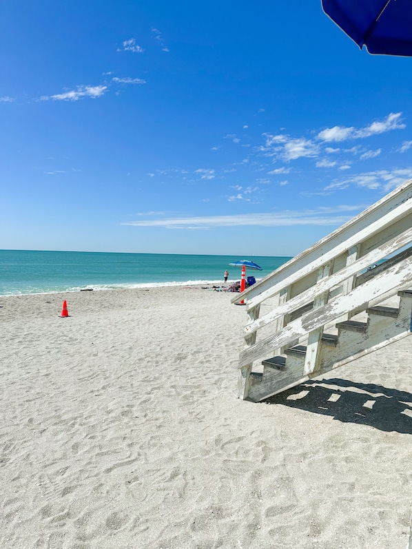 Een strand met een trap die naar het zand leidt, een blauwe parasol en een rode kegel.