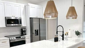 A modern kitchen with white cabinetry, stainless steel appliances, a large island with bar stools, and pendant lighting. - New! Beautiful Beach House, Venice (Venice)