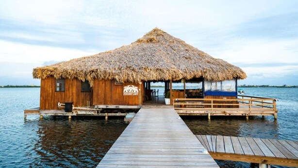 A wooden overwater bungalow with a thatched roof, a dock, and a sign reading "Coconut Cafe". - Brahma Blue Belize Sunset suite #7 (2Bed/2Bath) (Ambergris Caye)