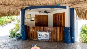 A thatched-roof veranda with a wooden reception desk, framed pictures, and a small table with books and a drink. - Brahma Blue Belize Sunset suite #7 (2Bed/2Bath) (Ambergris Caye)