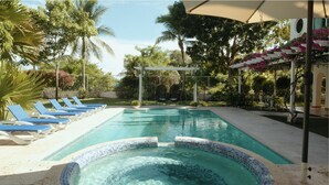 En swimmingpool med liggestole, et pergola overdækket med blomster og frodig vegetation omkring.