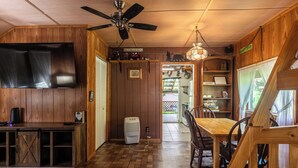A wooden interior with a ceiling fan, a flat-screen TV, a dining table with chairs, and a kitchen area visible through an open door.