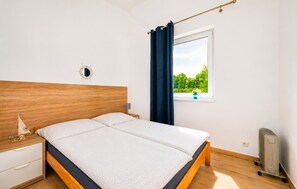 A bedroom with a wooden headboard, a bed with a white mattress, a small bedside table with a lamp, a window with a view of greenery, and a radiator. - Nice home in Jaroslawiec with WiFi (Jaroslawiec)