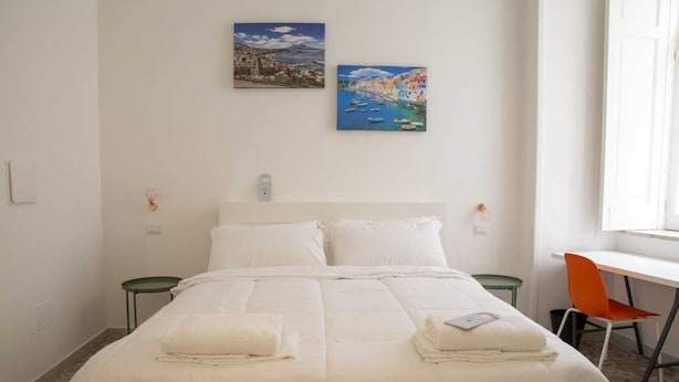 Habitación de hotel con una cama grande, dos almohadas, una mesita de noche, una silla y dos cuadros en la pared.
