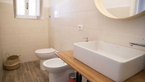 Un baño con una bañera blanca, un inodoro y un bidé. Hay una ventana con persianas, una encimera de madera y un espejo redondo sobre el lavabo.