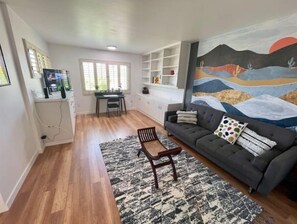 A living room with a sofa, a coffee table, and a rug. There is a painting on the wall and a window with blinds.