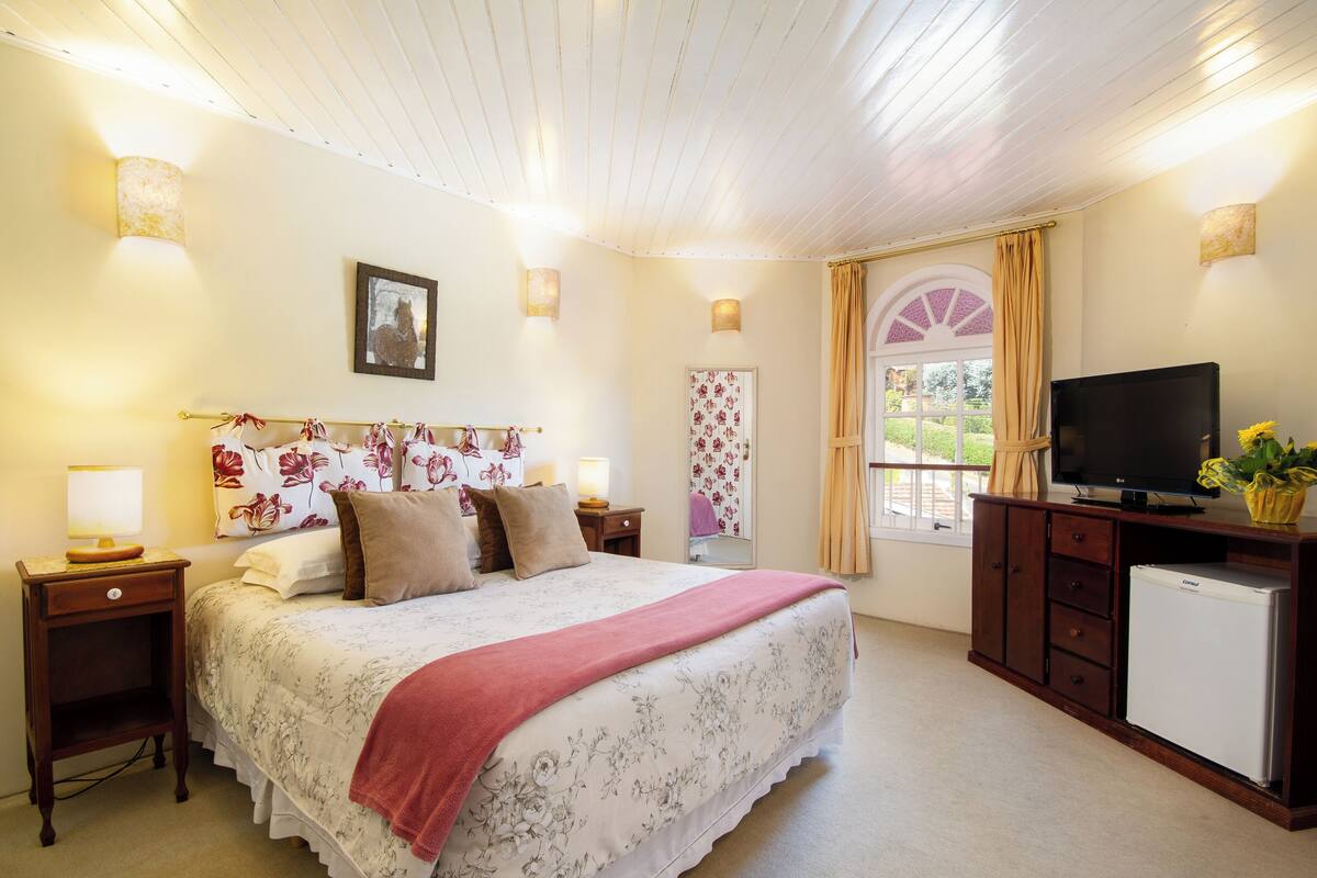 Un dormitorio con una cama grande, televisión, un refrigerador pequeño y una ventana con cortinas.