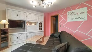 Un salon avec un canapé en cuir noir, une table basse en bois et un téléviseur posé sur un meuble blanc. Un mur rose orné d’une citation de Disney est visible.