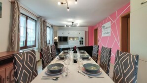 Un espace repas avec une longue table dressée pour six personnes, des chaises aux motifs géométriques, un lustre et une cuisine en arrière-plan.