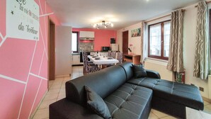 Un salon avec un canapé en cuir noir, une table à manger avec des chaises, une cuisine en arrière-plan et une fenêtre avec des rideaux.