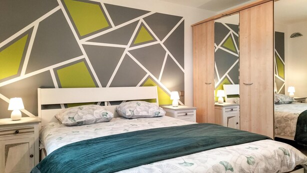 Une chambre à coucher moderne avec un mur orné d’un motif géométrique, un lit avec une literie à motifs floraux et une table de chevet avec une lampe.