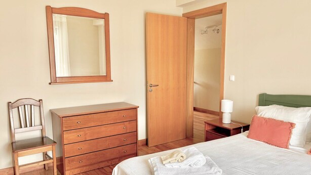 A bedroom with a bed, a wooden dresser, a chair, a nightstand with a lamp, and a door leading to a bathroom. - Casa dos Avós - Garden & Lake II (Ponta Delgada)