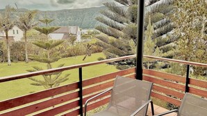 A balcony with a wooden railing and two mesh chairs, overlooking a landscaped garden with various trees and a distant view of houses and hills. - Casa dos Avós - Garden & Lake II (Ponta Delgada)