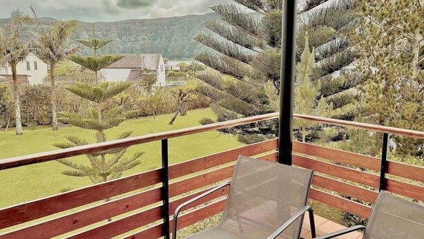 A balcony with a wooden railing and two mesh chairs, overlooking a landscaped garden with various trees and a distant view of houses and hills. - Casa dos Avós - Garden & Lake II (Ponta Delgada)