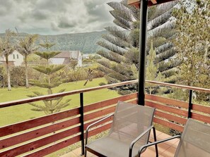 Un balcón con barandilla de madera y dos sillas de malla, con vista a un jardín ajardinado con varios árboles y una lejana vista de casas y colinas.