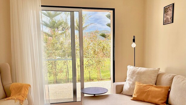 A living room with a sofa, a small round table, and a view through a sliding glass door to an outdoor area with trees and a clear sky. - Casa dos Netos - Garden & Lake III (Ponta Delgada)