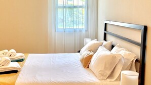 A neatly made bed with white linens and a wooden headboard, a window with sheer curtains, and a small table with a lamp and towels. - Casa dos Netos - Garden & Lake III (Ponta Delgada)