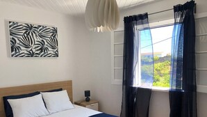 A neatly made bed with white and navy bedding, a bedside table with a lamp, a wall-mounted artwork, and sheer curtains over a window with a view of greenery. - A house in Caloura - Blue & Beach (Caloura)