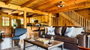 Interior acolhedor de uma cabana com uma escada de madeira, uma área de jantar com mesa posta para dois e uma sala de estar com sofá de couro e mesa de centro.