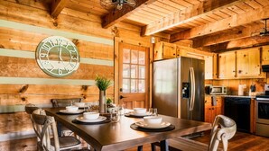 Interior de cabana de madeira com mesa de jantar posta para quatro pessoas, um relógio de parede grande e área de cozinha com eletrodomésticos modernos.