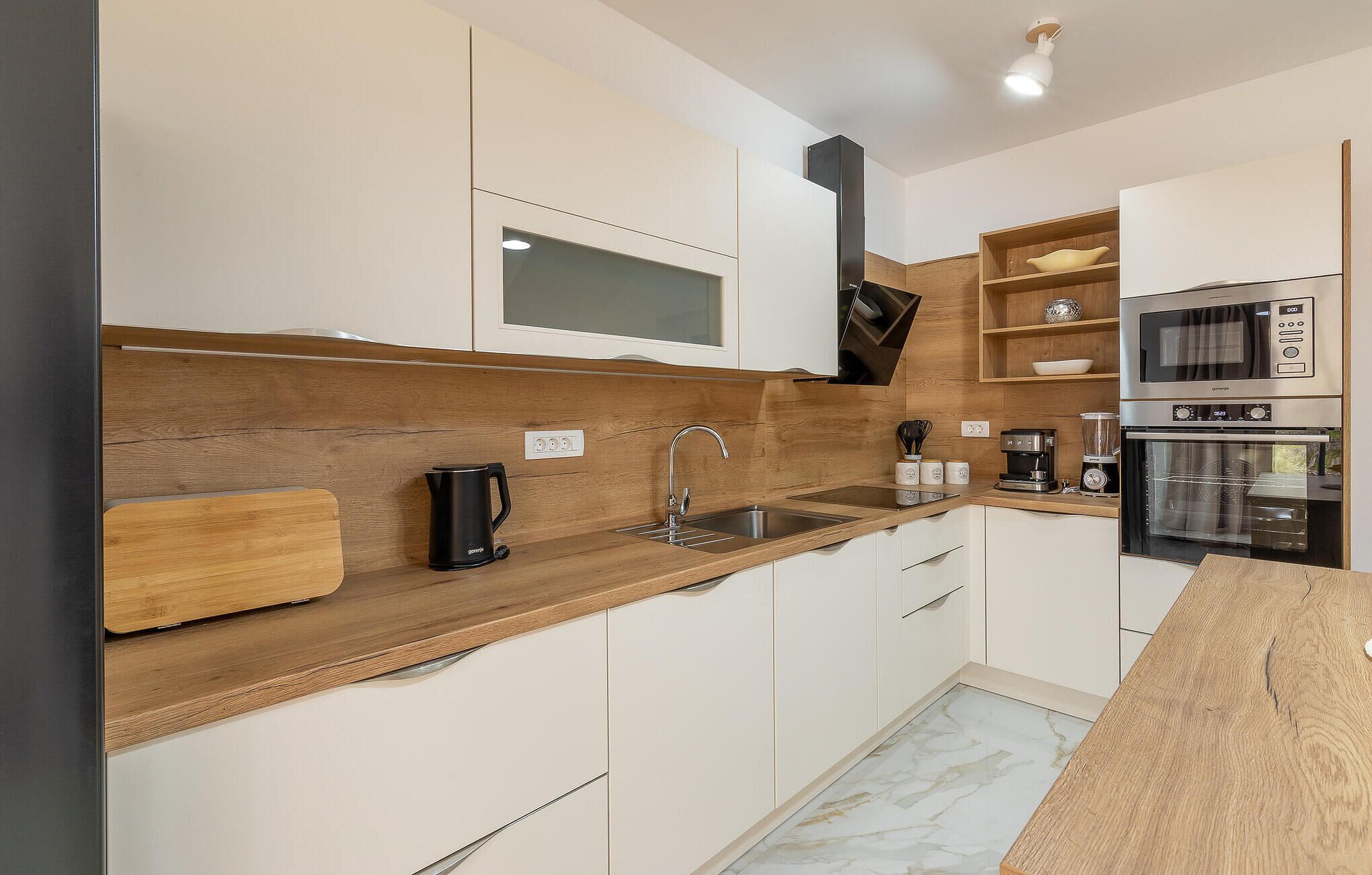 A modern kitchen with white cabinets, a wooden countertop, a coffee maker, a kettle, and a built-in oven.