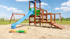 Eine Holzspielanlage mit einer blauen Rutsche und einer grünen Kletterwand, auf sandigem Untergrund mit Spielzeug in der Nähe.
