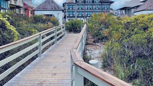 A wooden boardwalk leading to a large, multi-story building with a tower, surrounded by lush vegetation and a clear blue sky. - Luxury Beach House: Rooftop Jacuzzi & Family Fun (Inlet Beach)