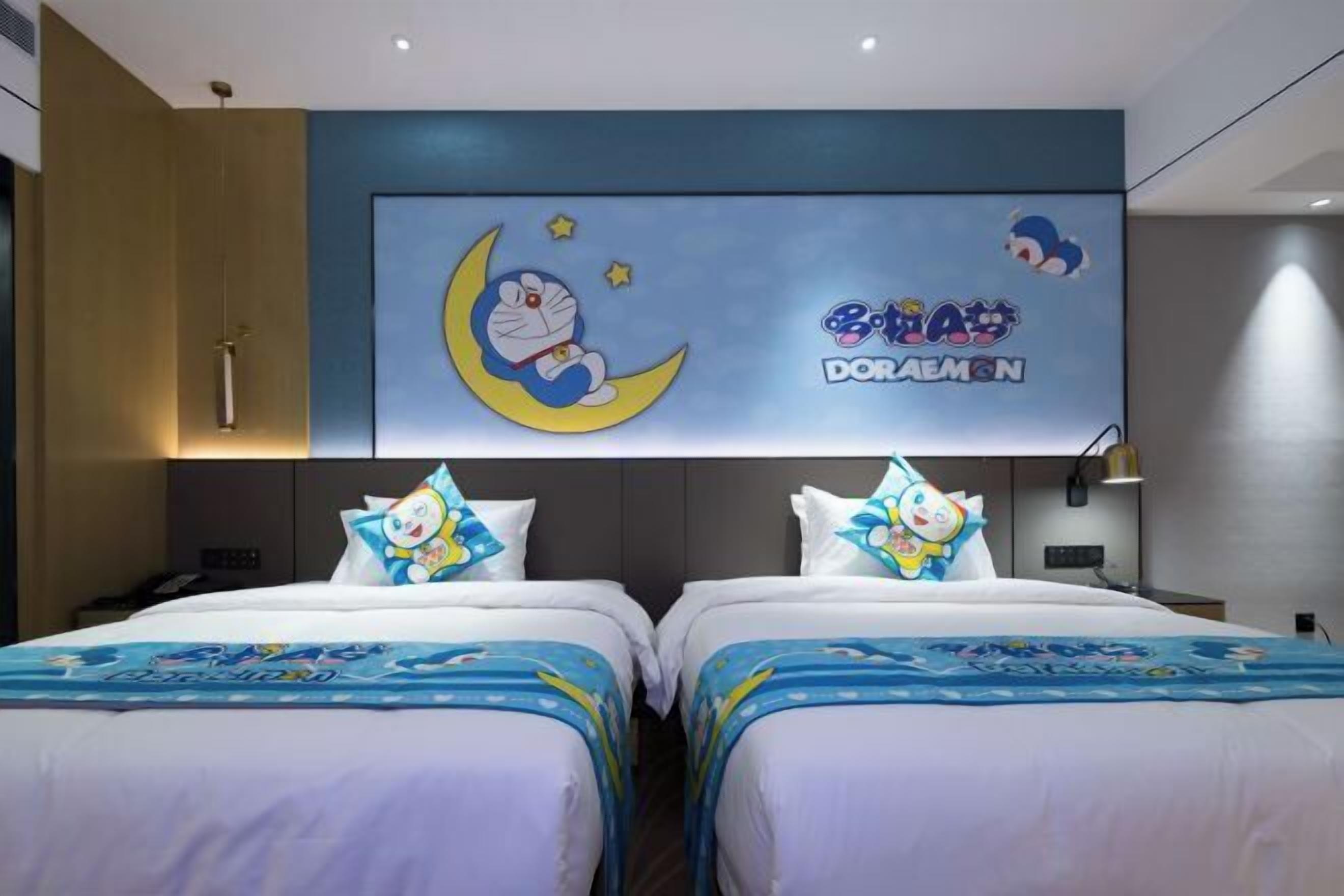 Ein Hotelzimmer mit zwei Betten, jedes mit einem Doraemon-Motiv auf der Bettdecke und dem Kopfkissen. An der Wand über den Betten ist ein großes Wandbild mit Doraemon zu sehen.