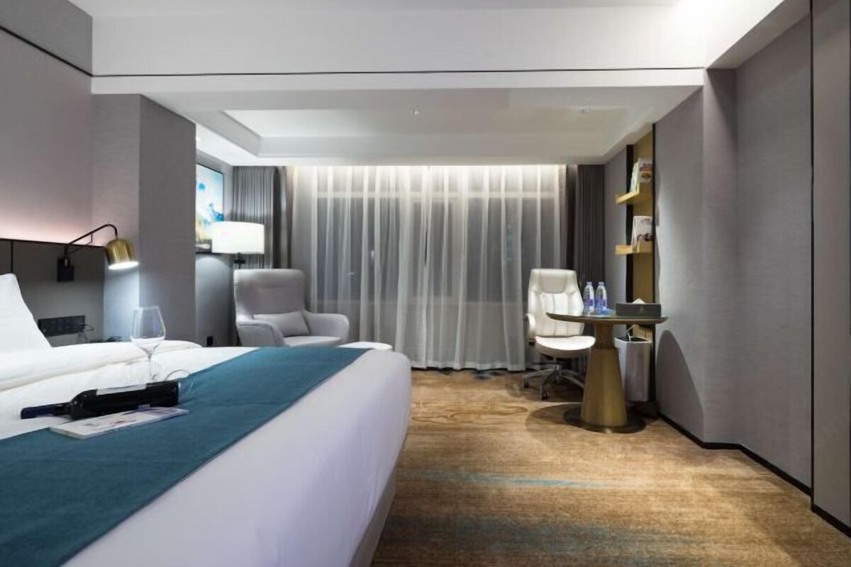 A modern hotel room with a large bed, a desk with a chair, a lamp, and a view of the city through the window.