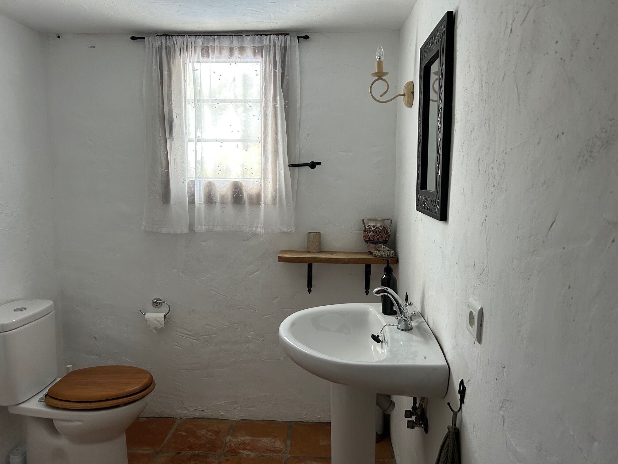 A small, white-walled bathroom with a wooden toilet, a white sink, and a window with sheer curtains.